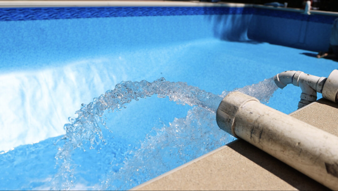 Landscape photo of two pipes shooting water into an empty vinyl liner pool. The photo is taken at a low angle left of two pipes lying on concrete, slightly over the edge of the pool.