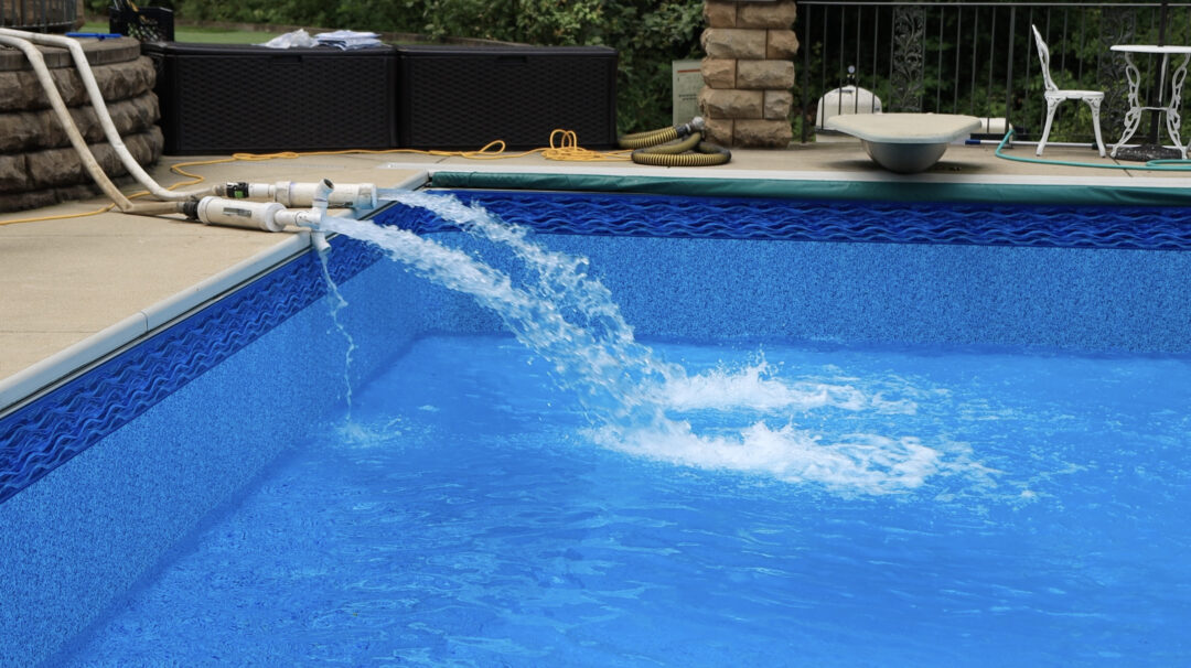 Landscape photo of the left-side deep-end corner of a vinyl liner pool. On the left are two pipes connected to hoses shooting water into the pool. In the background is a fence, two black containers, a white chair and table, a diving board overhanging the pool, and a fire pit cut off by the upper-left corner of the image.