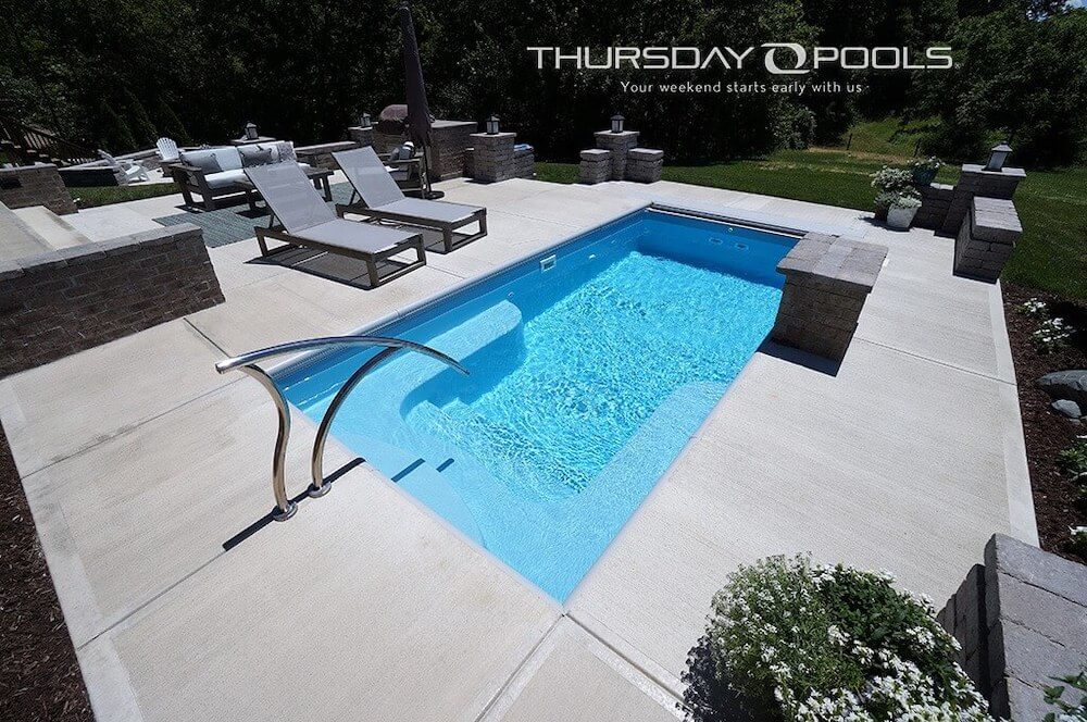 Large concrete patio backyard surrounded by trees with a light-blue inground fiberglass pool with brick retaining walls at each corner. A watermark says "THURSDAY POOLS Your weekend starts early with us" in the upper-right corner.