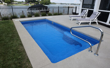 Landscape photo of a small, blue, rectangular, inground pool with a handrail surrounded by a small concrete patio off the back of a house. Beyond the pool is a fence with a large body of water behind it. On the far left-side of the image next to the patio is grass. A boat can be seen on the water just behind the pool and fence.