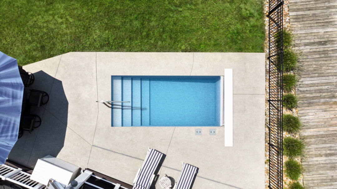 An aerial landscape photo of a portion of a backyard with a light blue rectangular inground pool in the center. The pool features an entry handrail and an automatic cover box. The pool is surrounded by a light gray concrete patio, atop which sit two lounge chairs next to the pool and an umbrella table area off the shallow end, cut off by the left-side edge of the image. Above the patio is grass, cut off by the top of the image. To the right of the patio is a fence, with wood flooring beyond that, cut off by the right-side edge of the image. 