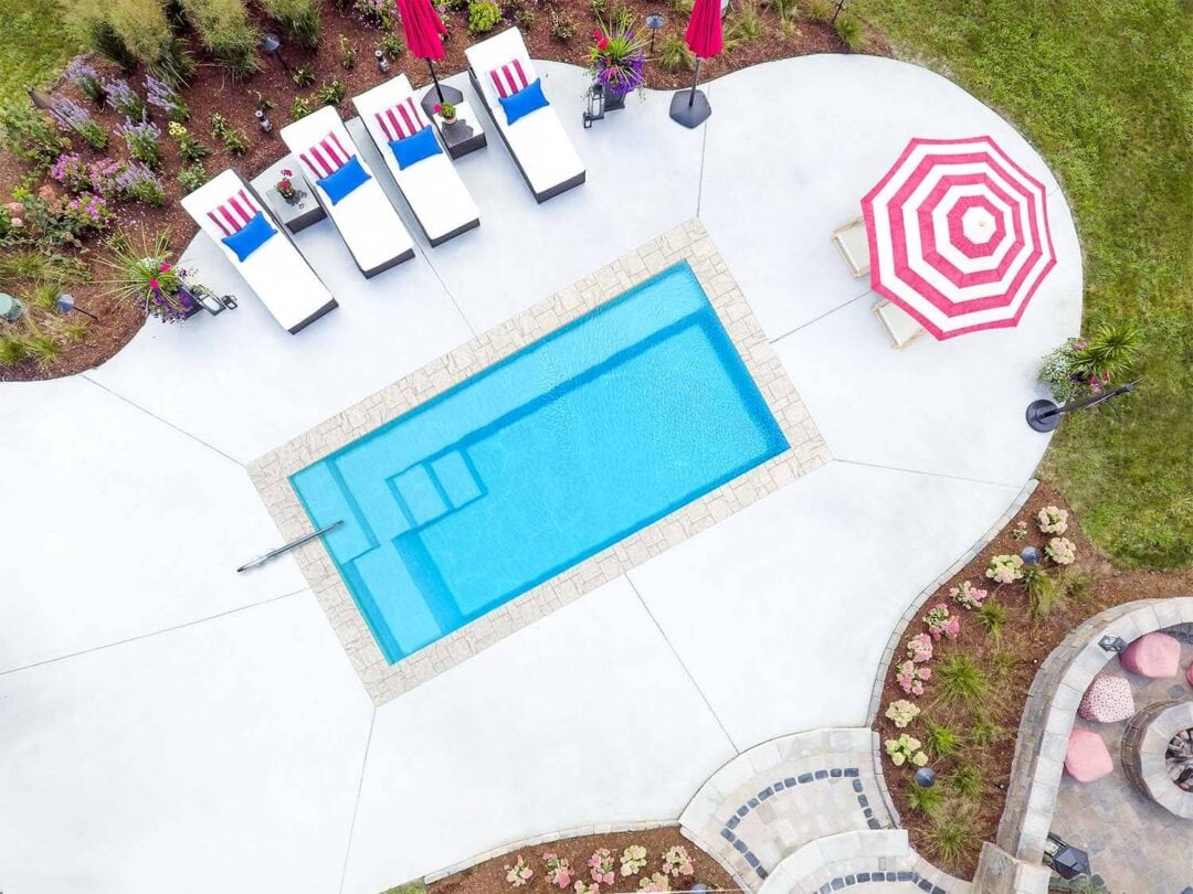 Aerial landscape photo of a rectangular light-blue inground pool featuring a handrail on the shallow-end width. The pool is surrounded by a light gray freeform concrete patio, atop which sit an umbrella off the deep-end width, and four lounge chairs parallel with the long side of the pool near the upper left corner of the image. The patio is partially surrounded by a garden at the top and bottom of the image. Beyond the mulch and concrete is grass. 