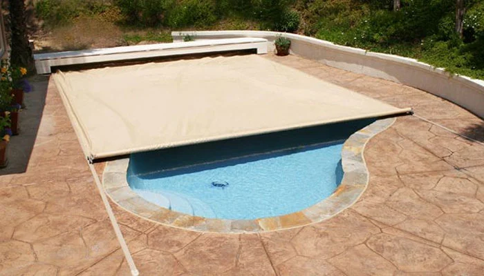 Landscape photo of an inground freeform pool featuring an automatic cover that is little over two-thirds closed. The pool is surrounded by a large stamped concrete deck.