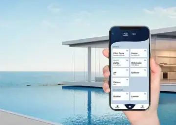 In the foreground right-side is a hand holding a phone that displays an app with pool feature buttons. In the background is an infinity pool overlooking a large body of water.
