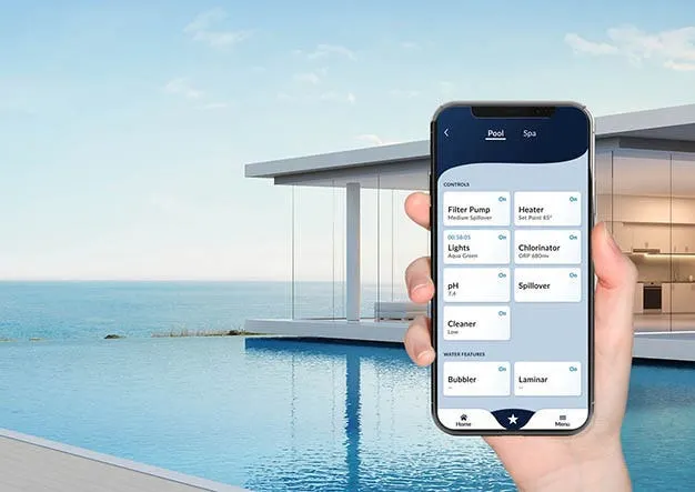 In the foreground right-side is a hand holding a phone that displays an app with pool feature buttons. In the background is an infinity pool overlooking a large body of water.