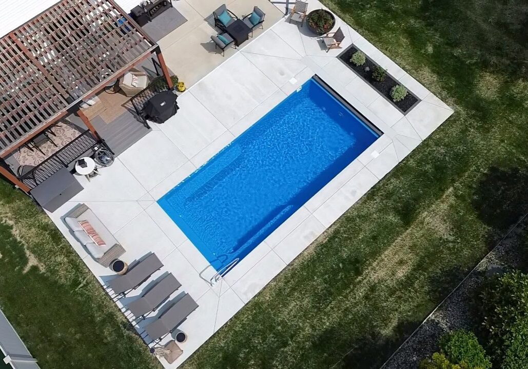 Aerial photo of a backyard featuring an inground pool surrounded by a concrete patio connected to the back of the house. In the top left corner off the house is a pergola. Next to the pergola, along the shallow end width of the pool are three lounge chairs.