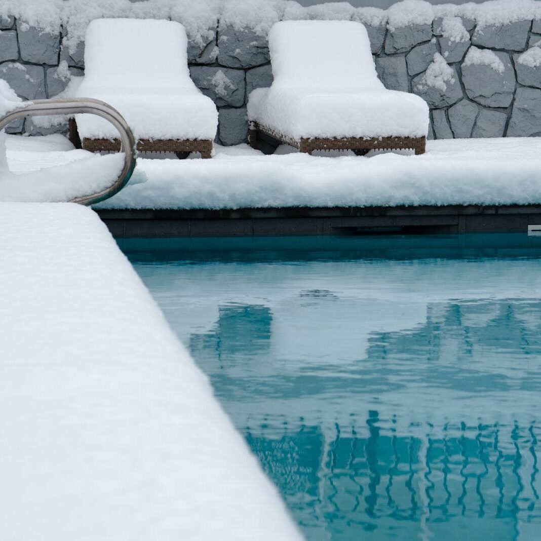 Inground pool with snow all around the pool. A brick wall and two lounge chairs can be seen in the background.