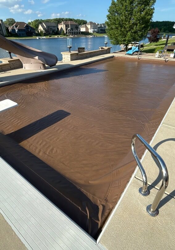 Closed inground pool with a brown automatic cover, white diving board, gray waterslide, a basketball hoop, and a fireplace surrounding the pool. a lake can be seen in the background.
