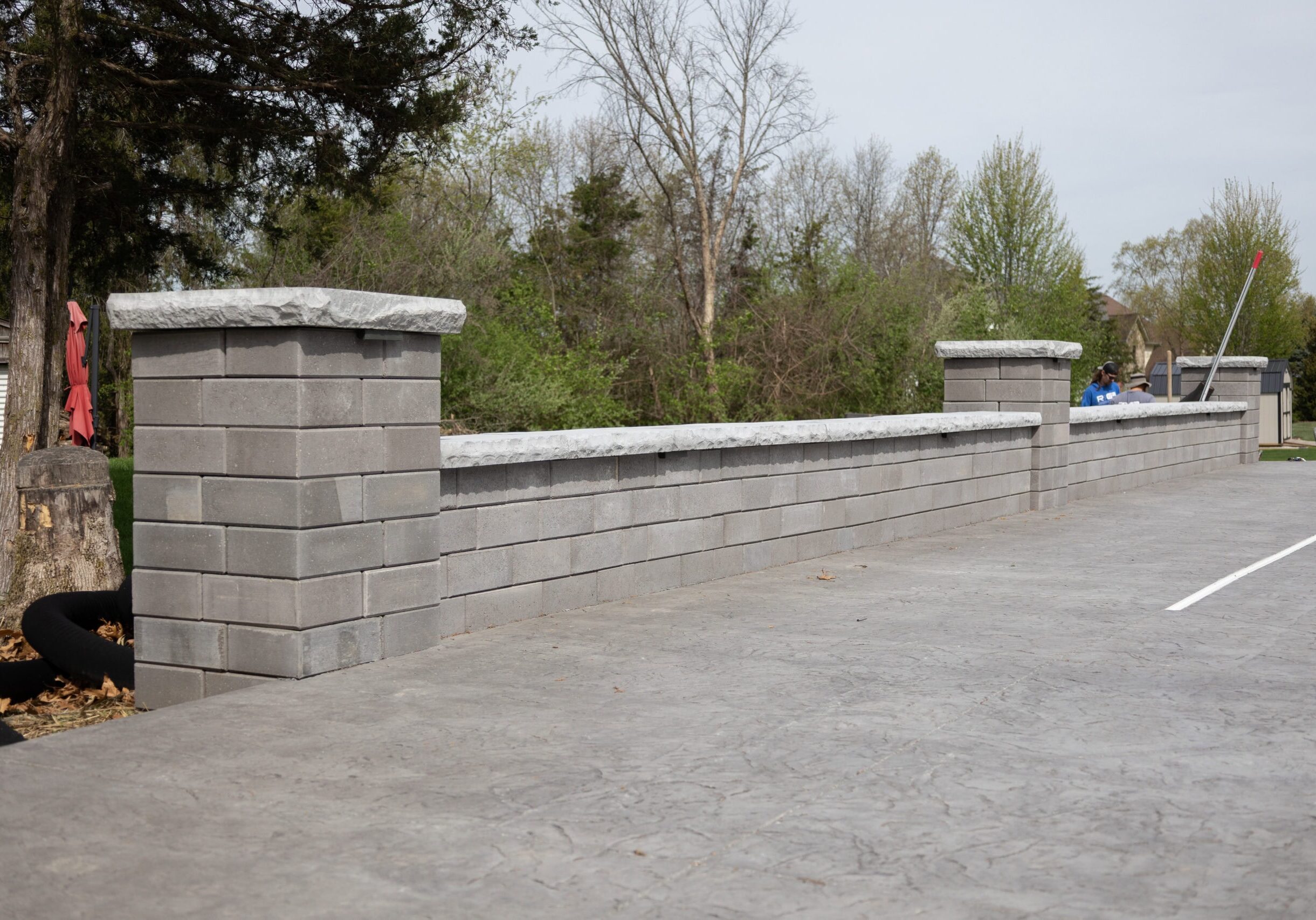 Landscape photo of a gray retaining wall propped up against a gray stamped concrete patio. The wall features three pillars, two on both ends and one in the middle. The retaining wall extends upward at an angle from the left side of the image to the right side. Trees can be seen in the background with the sky visible in the upper middle and right corner of the image.