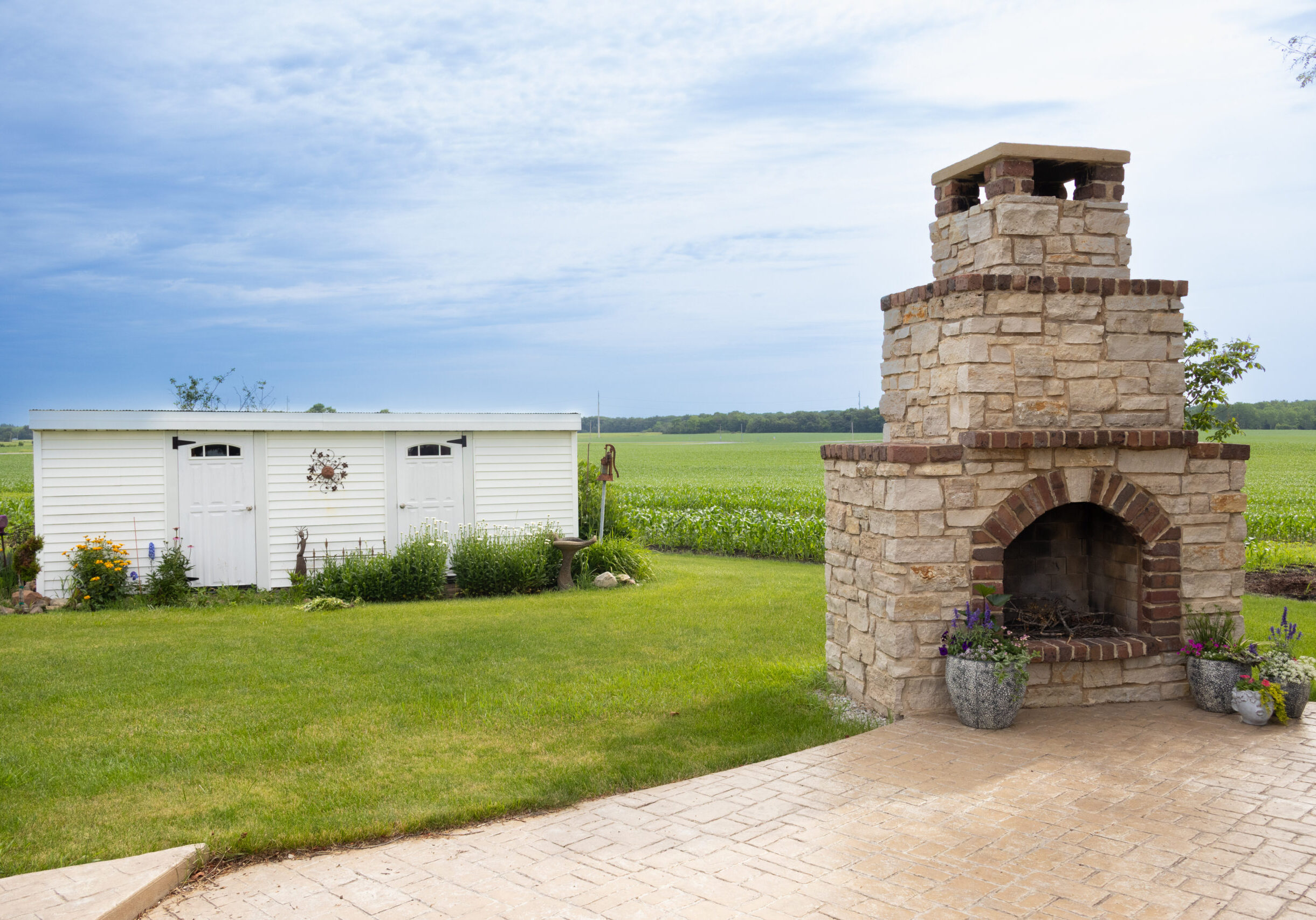 Photo of a fireplace off a concrete patio in a backyard. A wide white shed can be seen in the background.