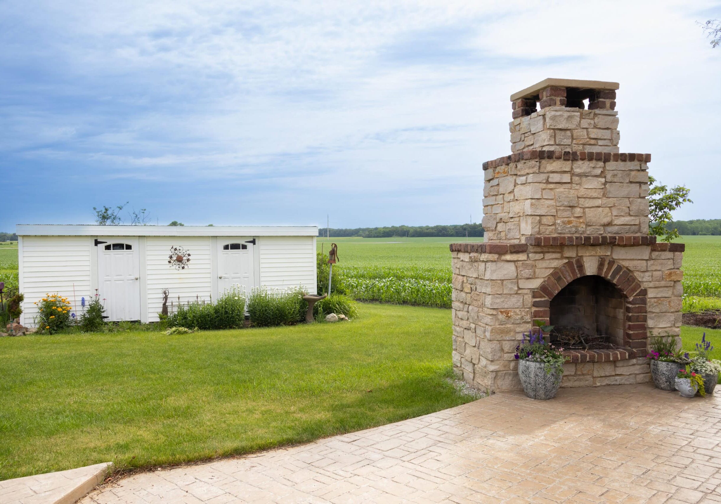 Photo of a fireplace off a concrete patio in a backyard. A wide white shed can be seen in the background.
