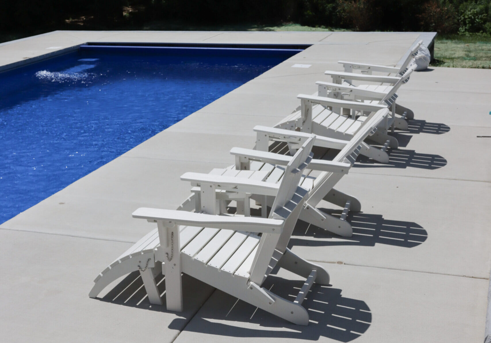 Photo of four white lounge chairs next to a blue inground swimming pool. The chairs are sitting on a concrete patio surrounding the pool.