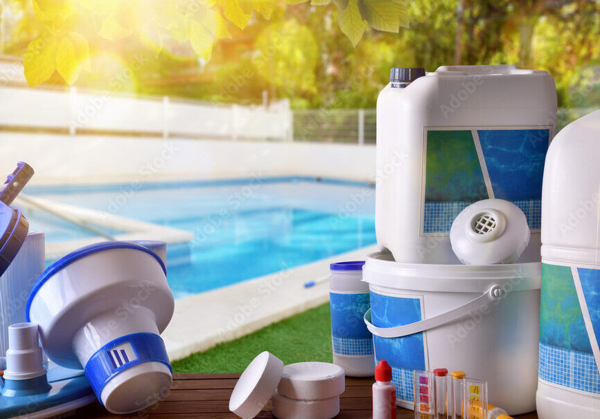 Photo of a variety of pool water chemicals sitting on a table in front of a light-blue inground swimming pool. Around the pool in the background is a white fence with trees behind it.