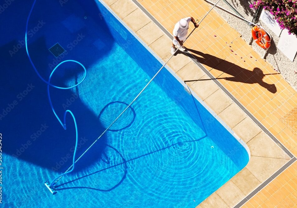 Aerial photo of a man vacuuming a blue inground swimming pool. The photo is only of a corner of the pool. The pool is surrounded by a rectangular brick paver border and a concrete patio.