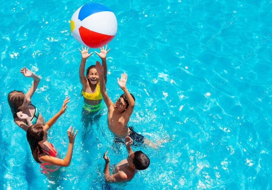 Aerial photo of five children bouncing a beach volleyball between one another in a swimming pool.