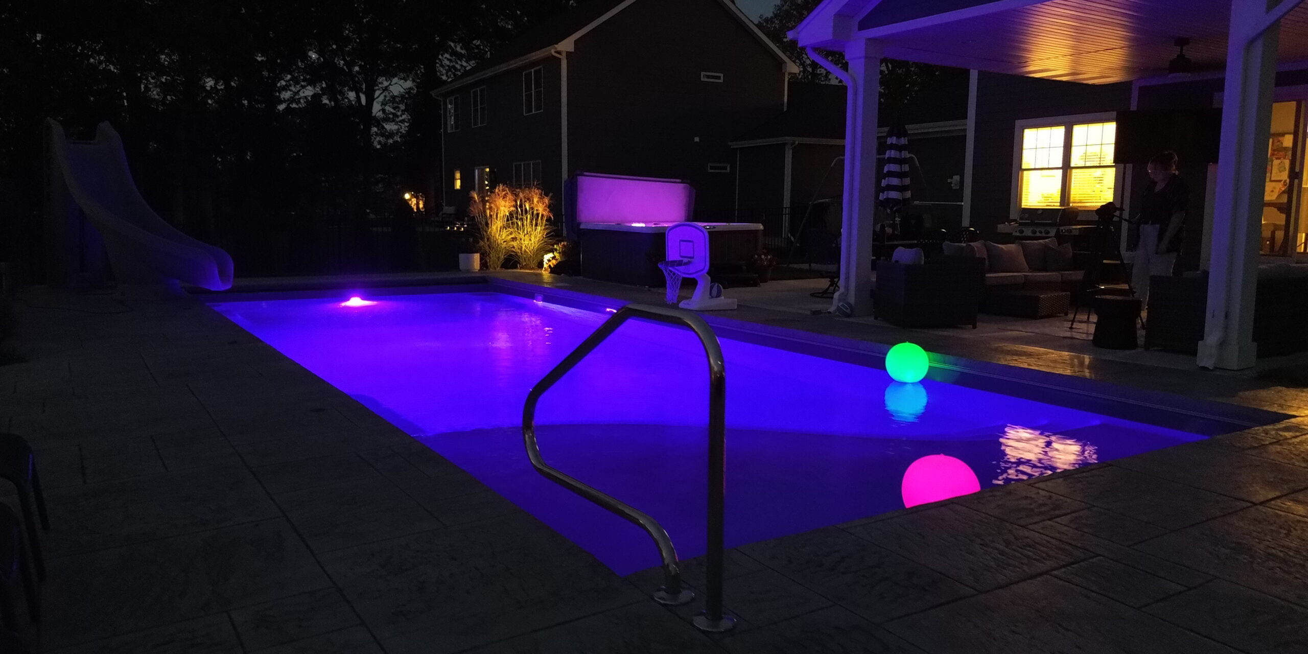 Inground pool with a purple pool light illuminating the water at nighttime. A glowing green and a glowing pink ball float in the pool. The pool features a slide on the left deep-end side, a basketball hoop, and a pool side gazebo. Next to the right-side deep end corner of the pool is an above ground hot tub also glowing purple.