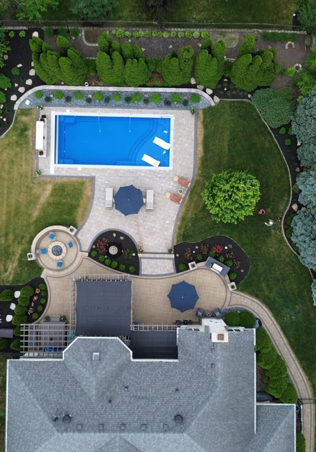 Portrait photo of a backyard featuring a curved concrete patio extending from the back of a house to a blue, rectangular, inground pool.