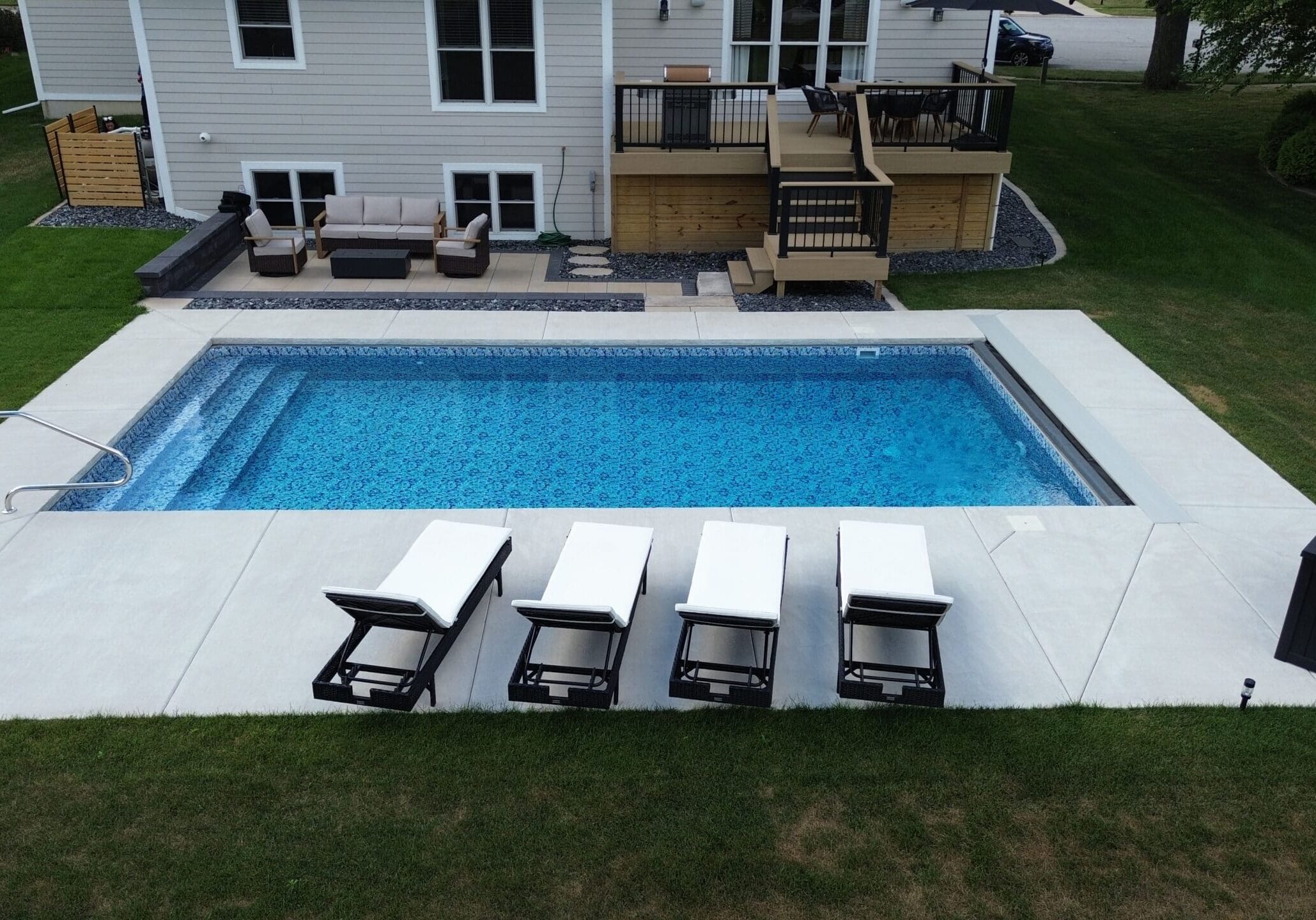 Landscape aerial photo taken of a light-blue, rectangular inground pool surrounded by a small, light-gray patio extending from a house which can be seen partially at the top of the image. Four black-and-white lounge chairs sit on the outside edge of the patio between the pool and grass that surrounds it. A black crate sits at the corner of the patio near the bottom right corner of the image.