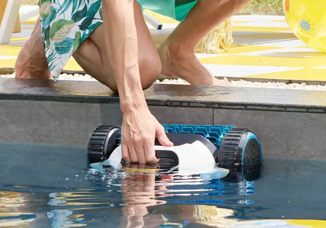 Person kneeled down outside an inground pool pulling a robotic pool cleaner out of the water from off of the pool wall.