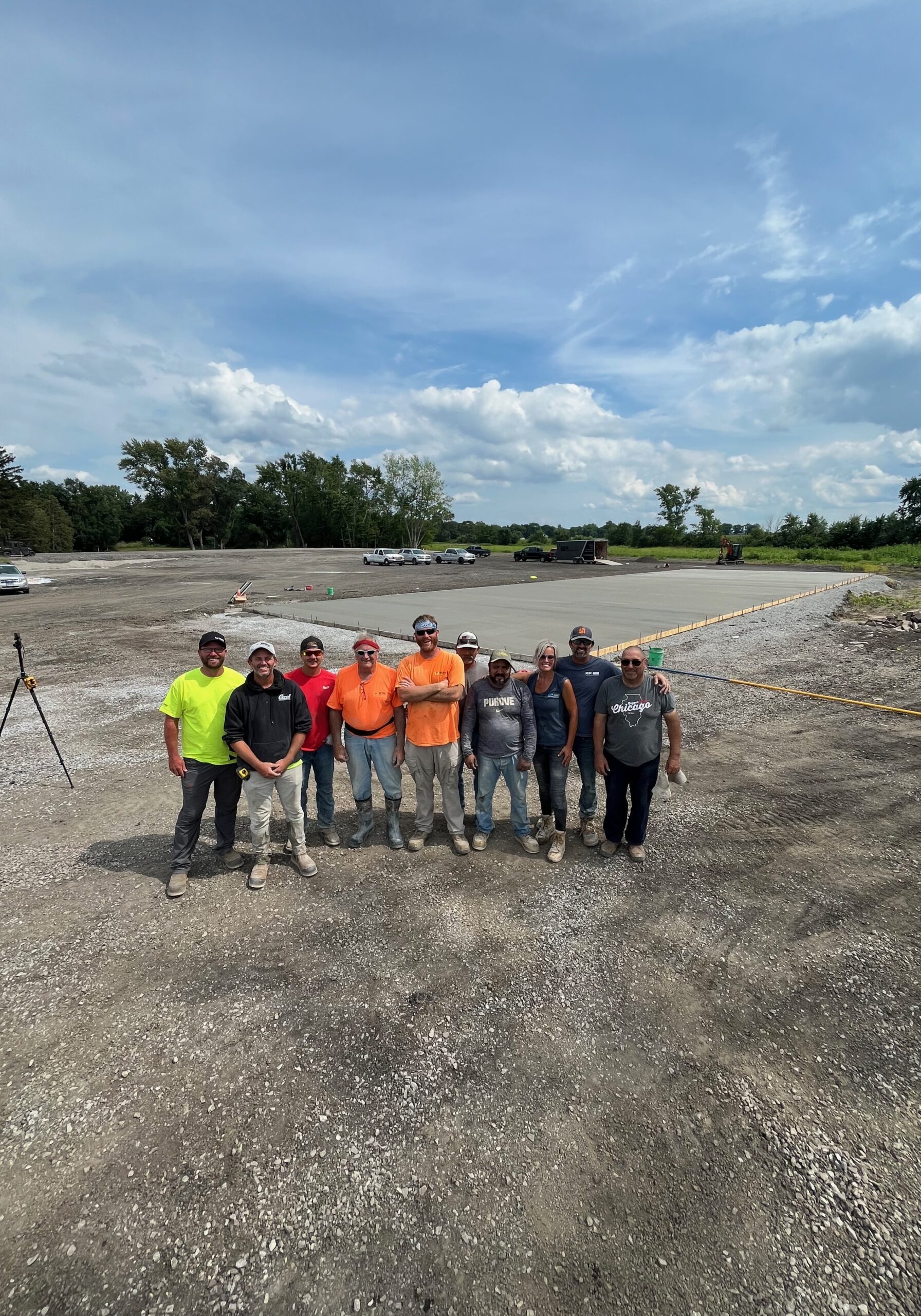 Portrait photo of nine men and one woman standing side by side center-frame on a large gravel ground with trees in the background. Behind the group is a large concrete slab that extends into the background.