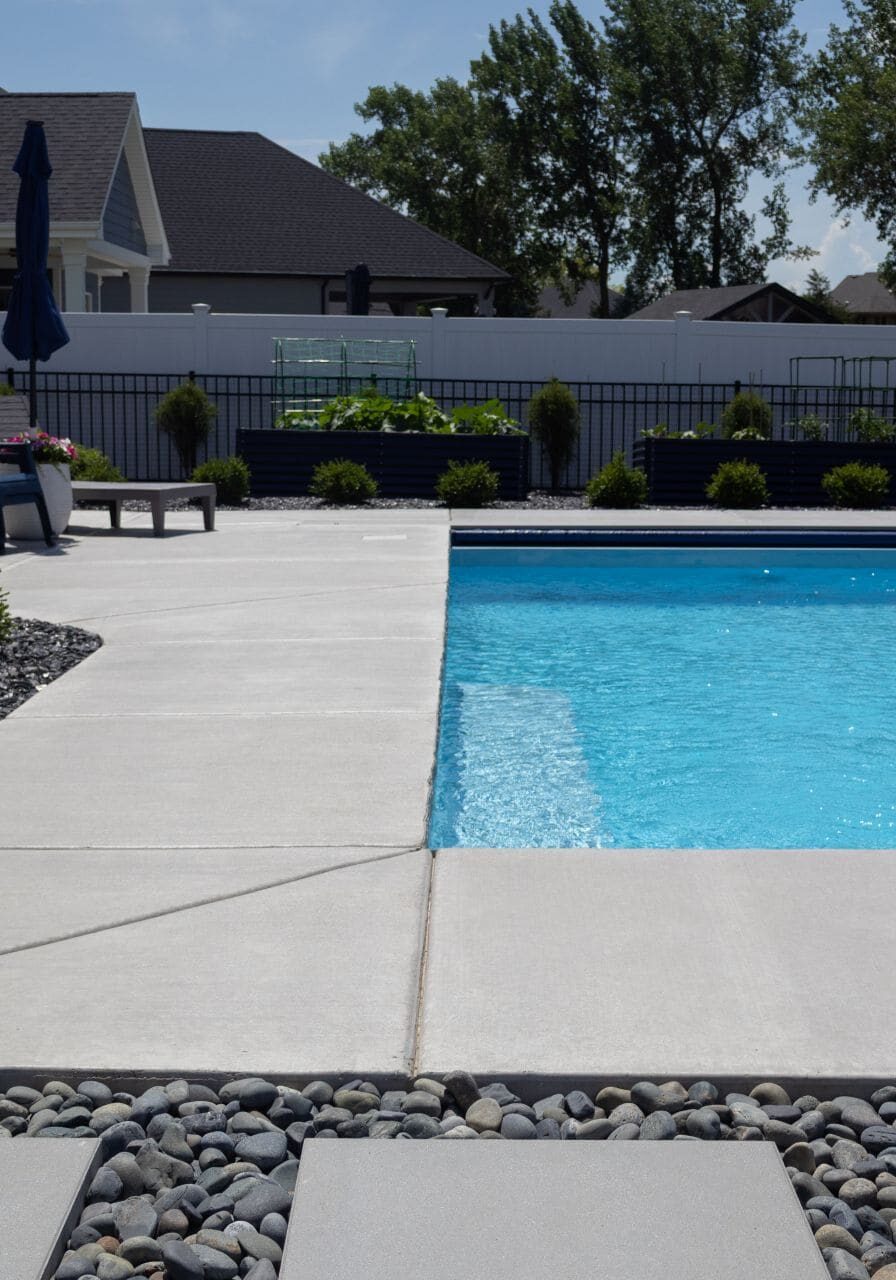 Light-blue rectangular inground swimming pool with a basketball hoop, surrounded by light-gray brushed concrete. In the foreground, outside of the concrete is black gravel and gray walkway stones. Shrubs and a white fence can be seen in the background behind the pool.