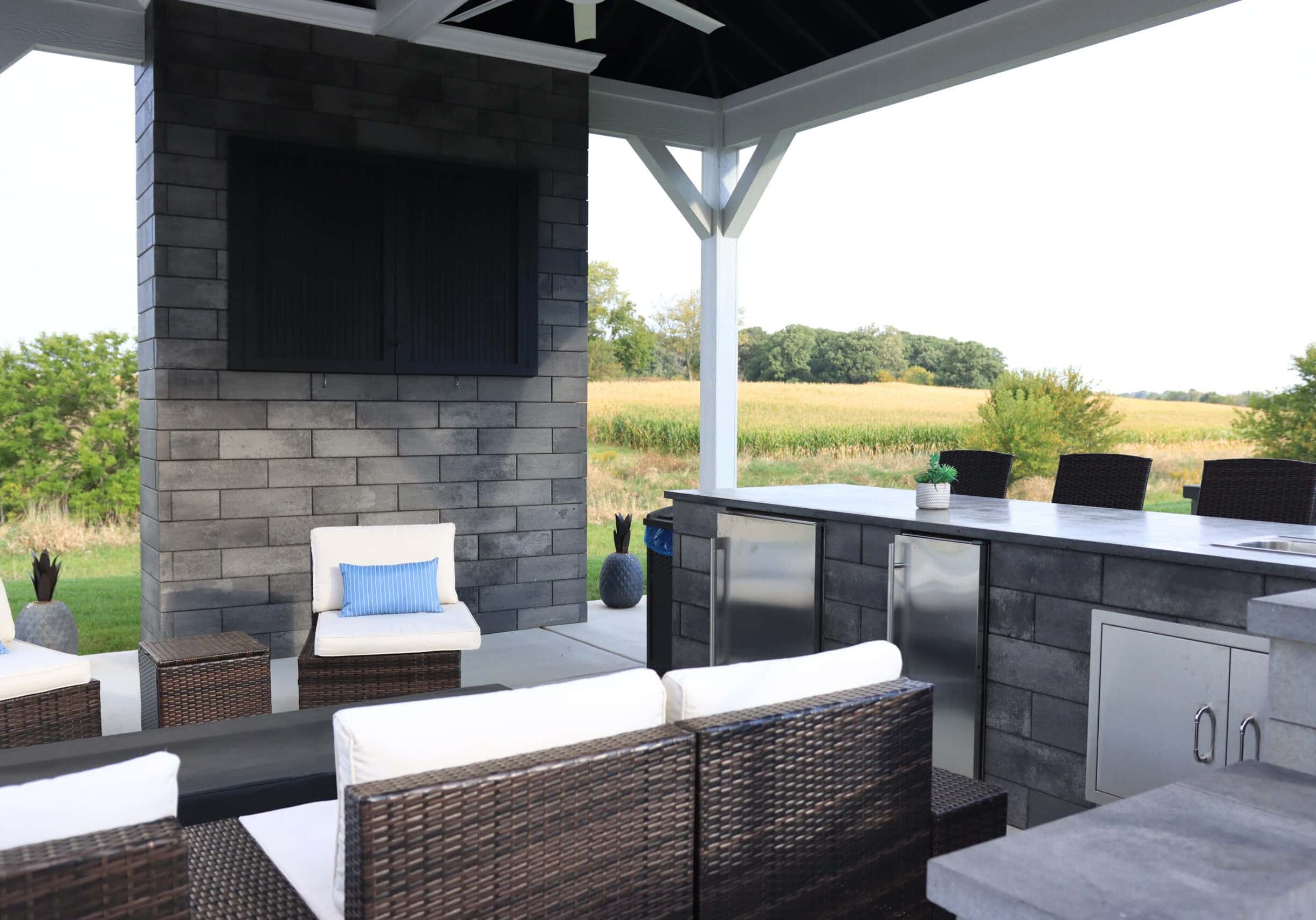 Landscape photo taken from underneath a gazebo of a large brick wall with a black rectangular housing at the top. Chairs sit in a semi-circle in front of the wall. A kitchen sits to the right of the chairs. A cornfield and trees can be seen in the background. The bottom of the roof can be seen cut off at the top of the image.