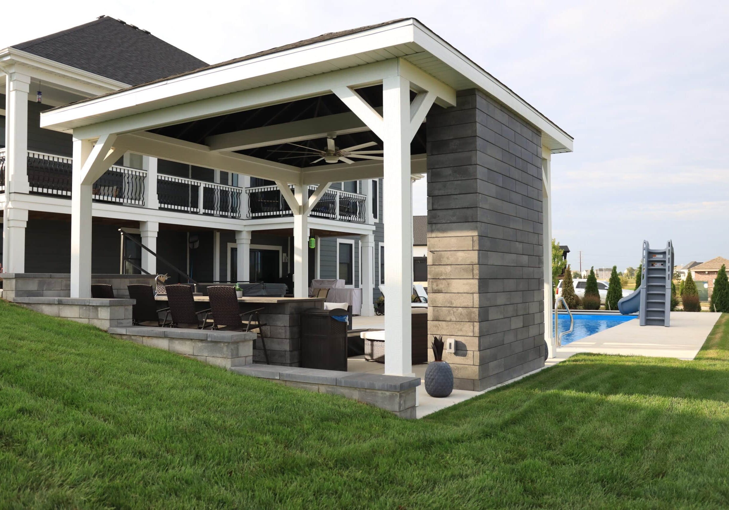Photo of the back of a gazebo in a backyard. Behind the gazebo you can see a small stretch of a light-blue inground pool. Behind the pool is a house.