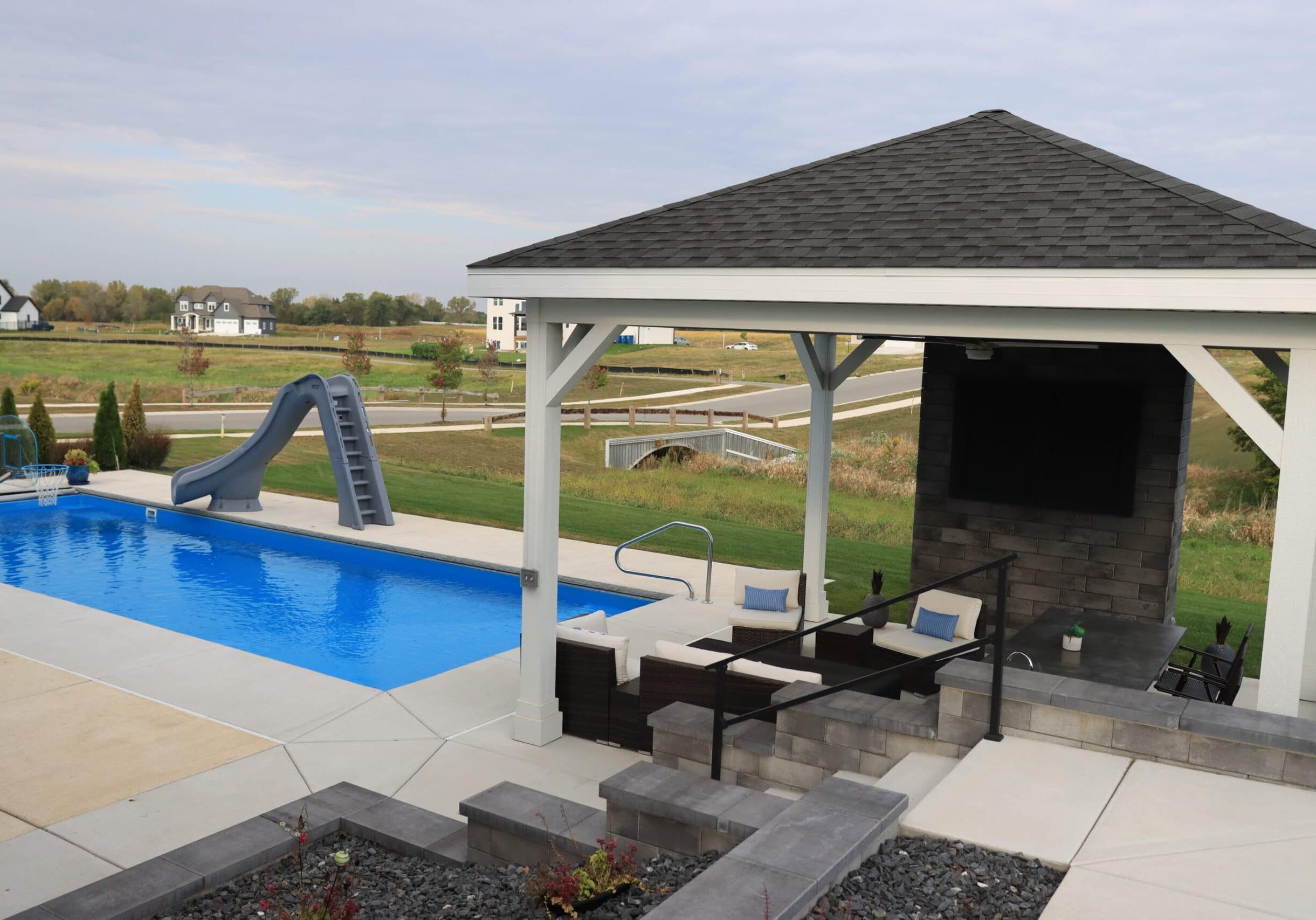 A backyard with a blue inground fiberglass pool surrounded by brushed concrete. Along the deep-end length is a pool slide. Along the shallow-end width is a gazebo.