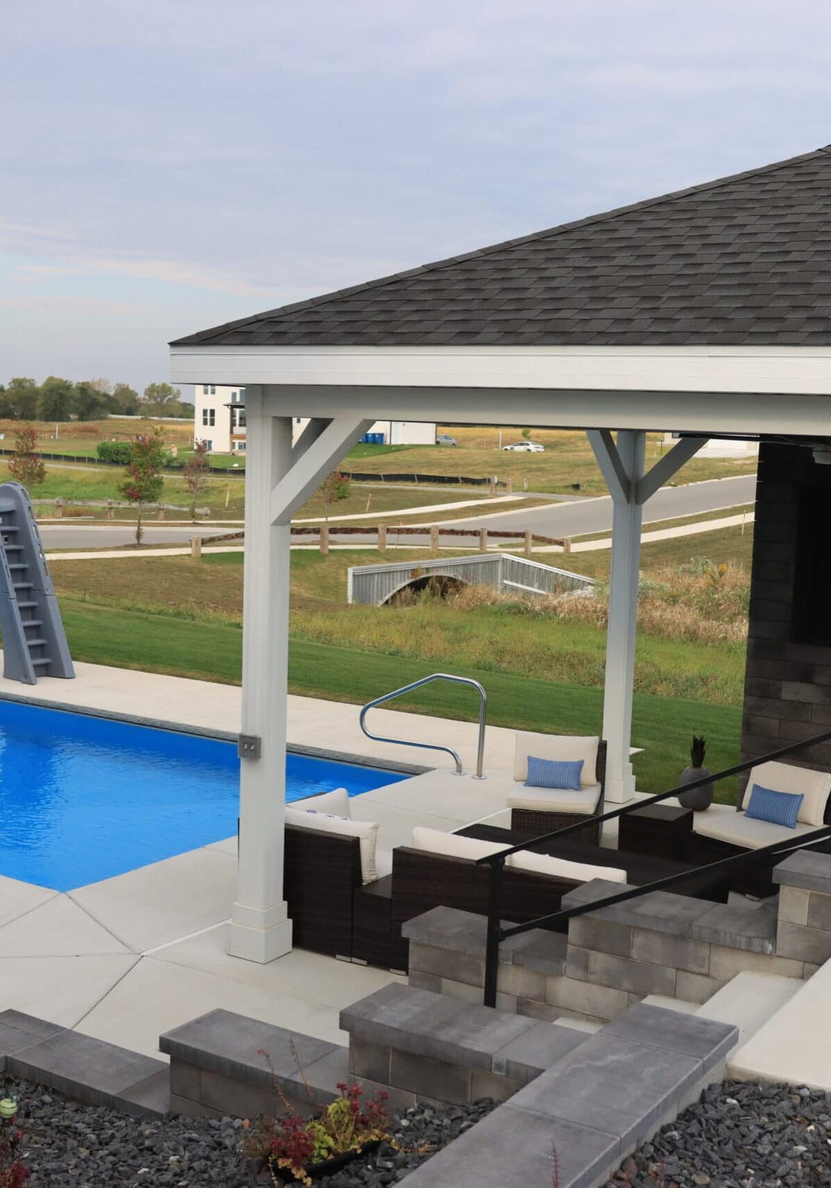 A backyard with a blue inground fiberglass pool surrounded by brushed concrete. Along the deep-end length is a pool slide. Along the shallow-end width is a gazebo.