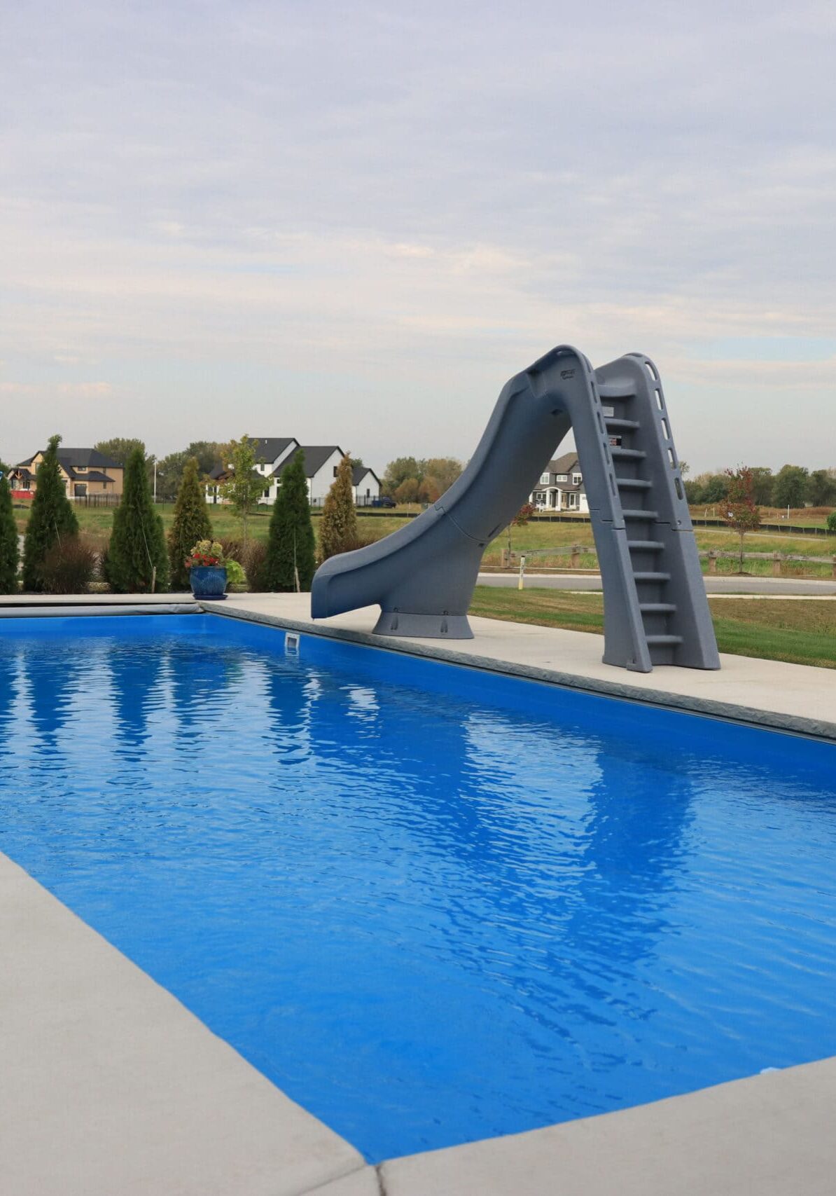 Inground fiberglass pool with slide and basketball hoop surrounded by a light-gray patio, with a row of trees across the deep end edge of the pool outside the patio.