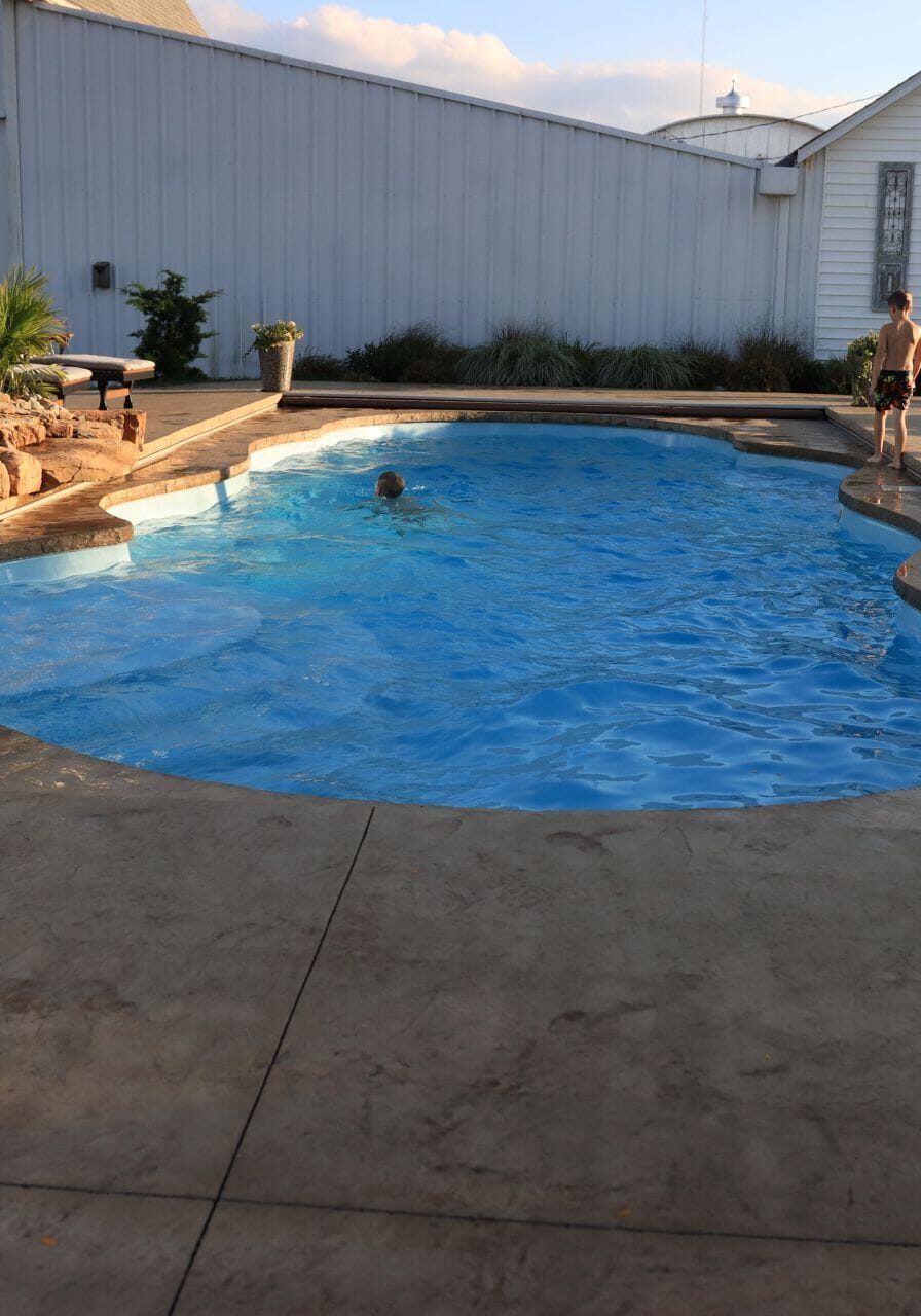 Photo of a freeform inground pool on a lowered deck surrounded by light-brown stamped concrete. Connected to the left side of the pool is a rock-waterfall. Behind the pool is a large white barn.