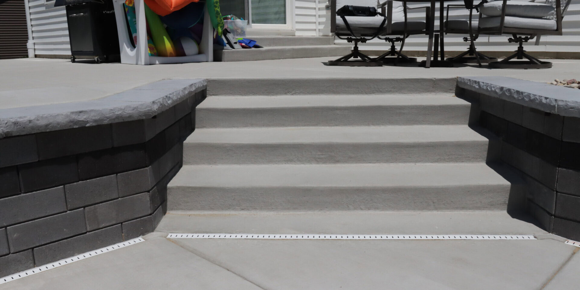 Landscape photo of a gray concrete staircase connecting an upper level of a concrete patio to a lower level. The staircase is slightly right of center in the image, with black brick pavers on each side. At the top of the image is the bottom of a light-gray house. On the left side of the image, propped up against the house is a crate full of floats and pool toys. On the other side of the stairs/image is a table with four rotating chairs.