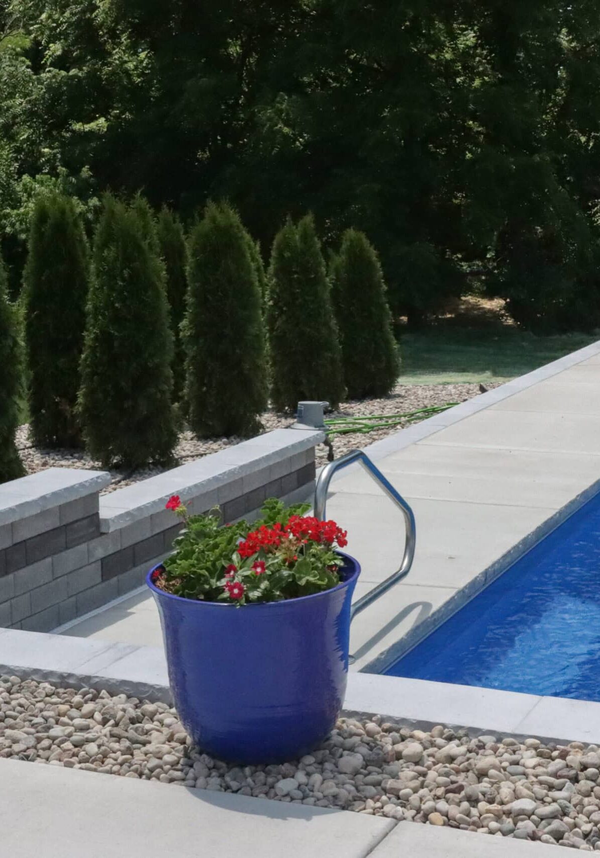 Landscape photo taken of a portion of a concrete pool patio in a backyard. The patio extends at an angle from the bottom left corner of the image to the upper right quarter of the image before extending right to the end of the image. At the bottom of the image is a thin rock bed against the edge of concrete that drops down to the pool patio via retaining wall on the left. A blue, rectangular inground pool can be seen on the left side of the image cut off by the edge. Small evergreen trees line the length side of the patio behind the retaining wall. The background is comprised of grass and tall, thick trees.