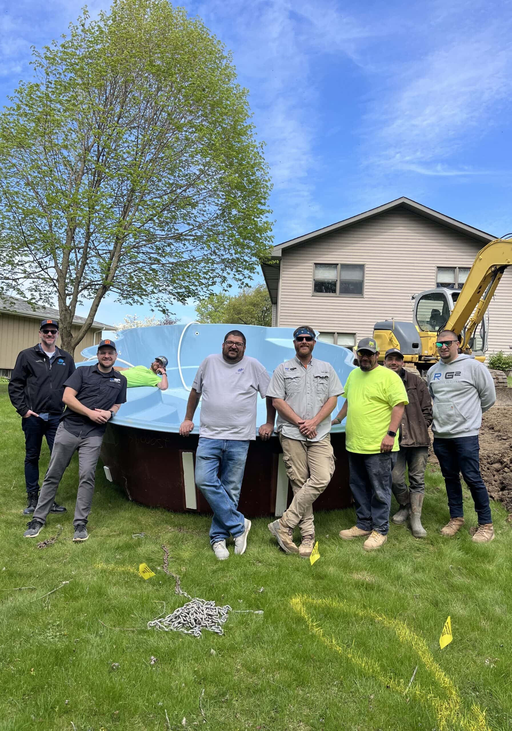 Fiberglass pool sitting on grass in a backyard next to a hole and guys standing in front of it.