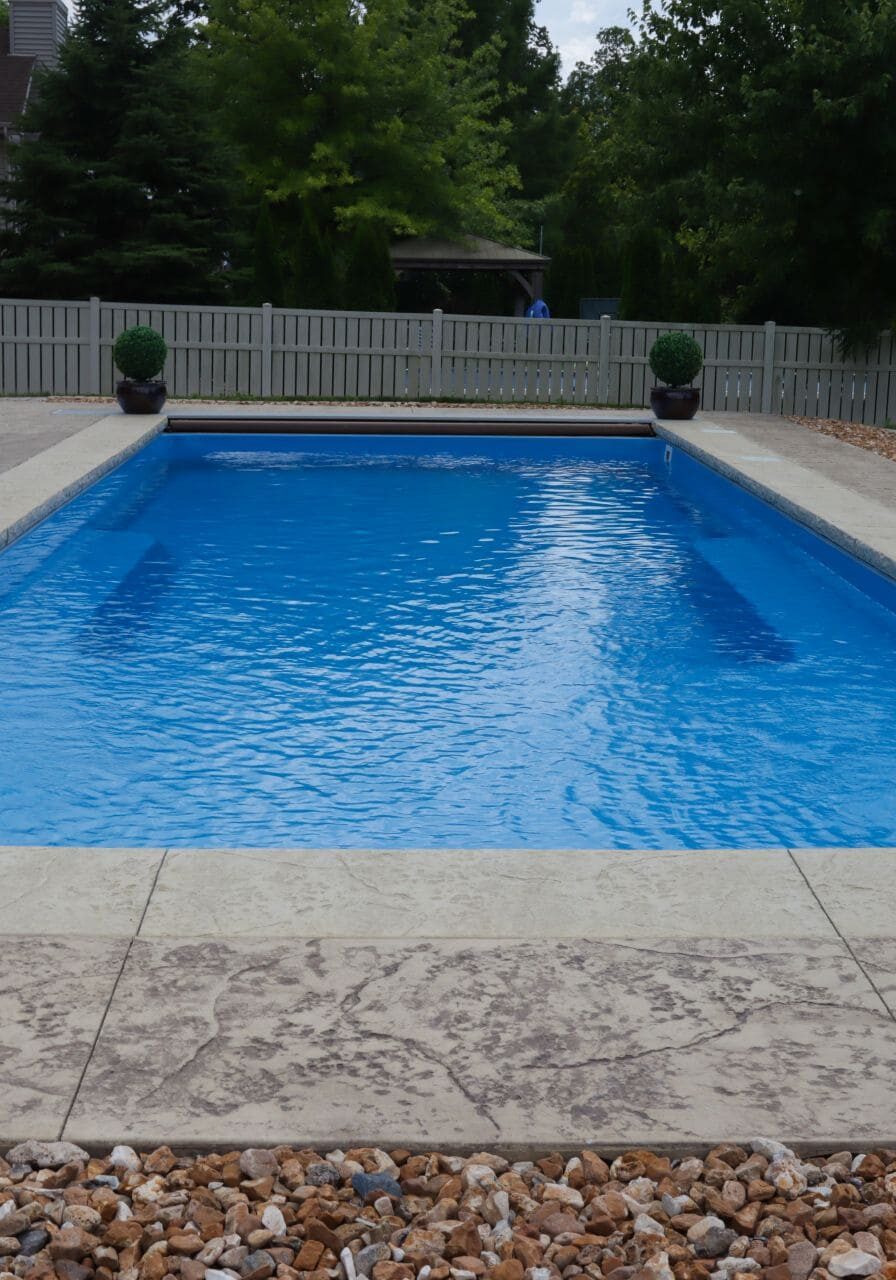 Photo of an inground pool surrounded by a stamped concrete pool deck with a few feet of gravel around the patio. Grass can be seen beyond gravel.