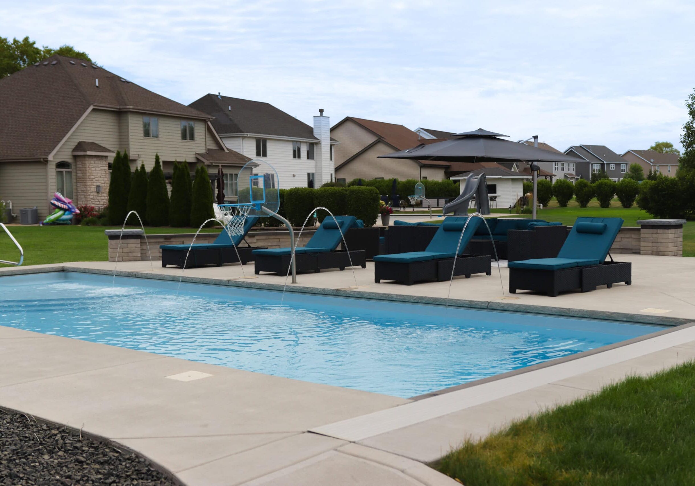 Landscape photo of a backyard featuring a large concrete patio surrounding a light blue, rectangular inground pool extending downward from the middle of the left-side edge of the image. Grass can be seen in the bottom right corner of the image. Behind the pool on the concrete sit four black-and-blue lounge chairs along the edge of the pools length. Additional furniture can be seen behind these chairs. Houses can be seen in the background extending from the left side of the image downward to the right.