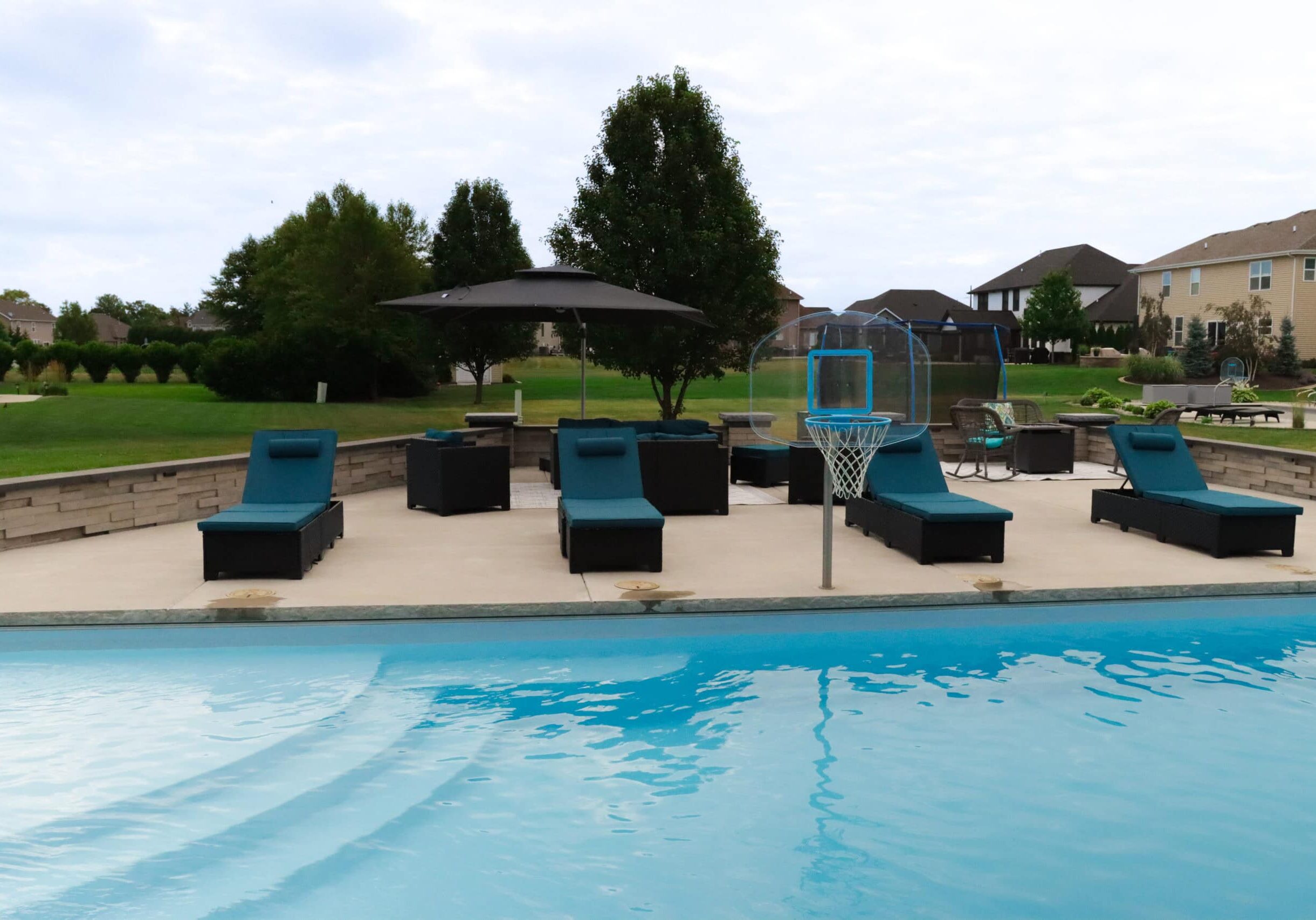 Photo of a backyard broomed concrete patio featuring a light-blue inground pool with a basketball hoop. Behind the pool on the patio are three lounge beach chairs. Behind the chairs are more small chairs, an umbrella, and a retaining wall surrounding the patio.