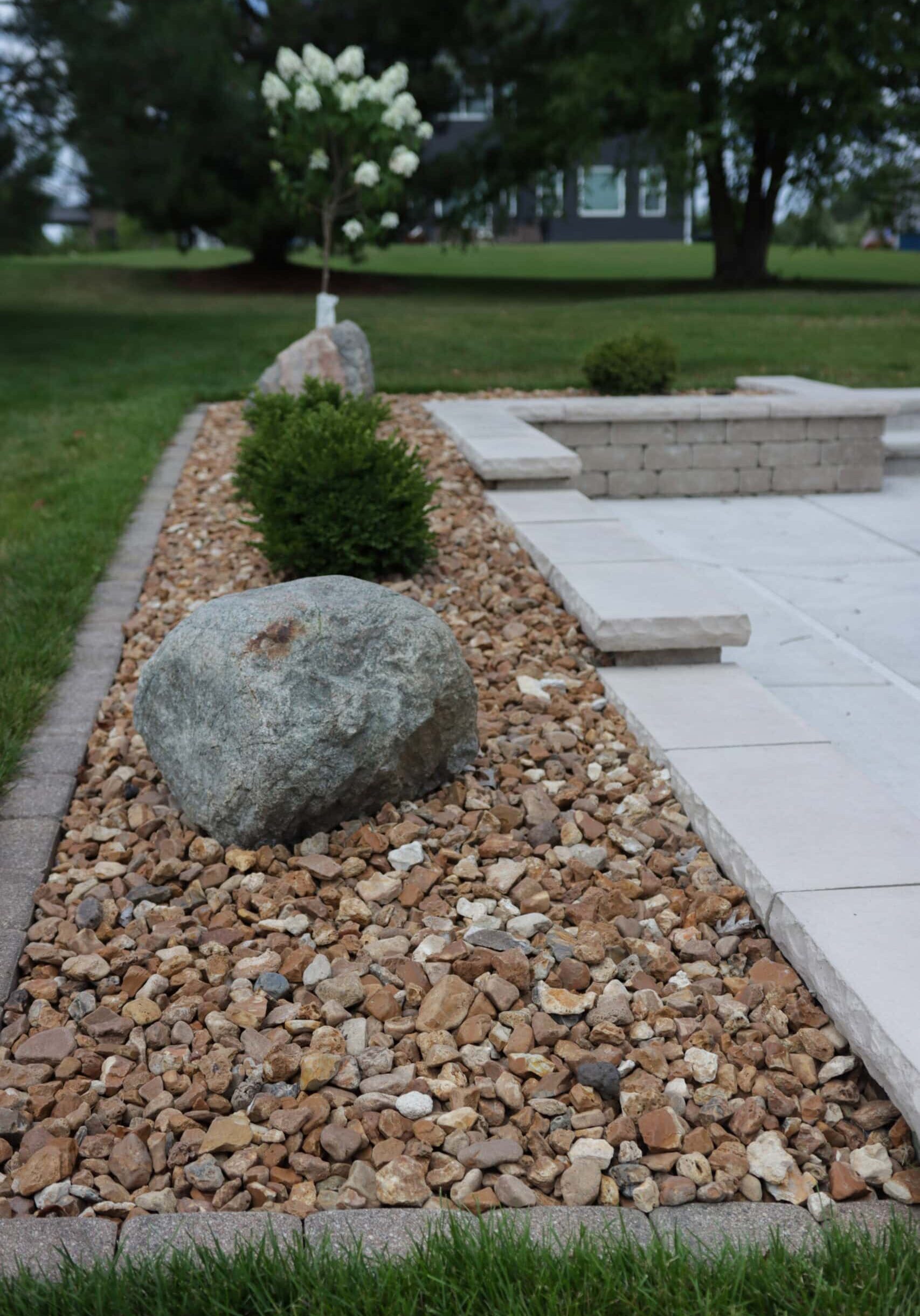 Portrait photo of the corner/edge of a concrete patio with a border rock bed. Atop the rocks sit two boulders and two plants between them. Grass can be seen at the bottom edge of the image and surrounding the concrete. Trees grow from the grass in the background.
