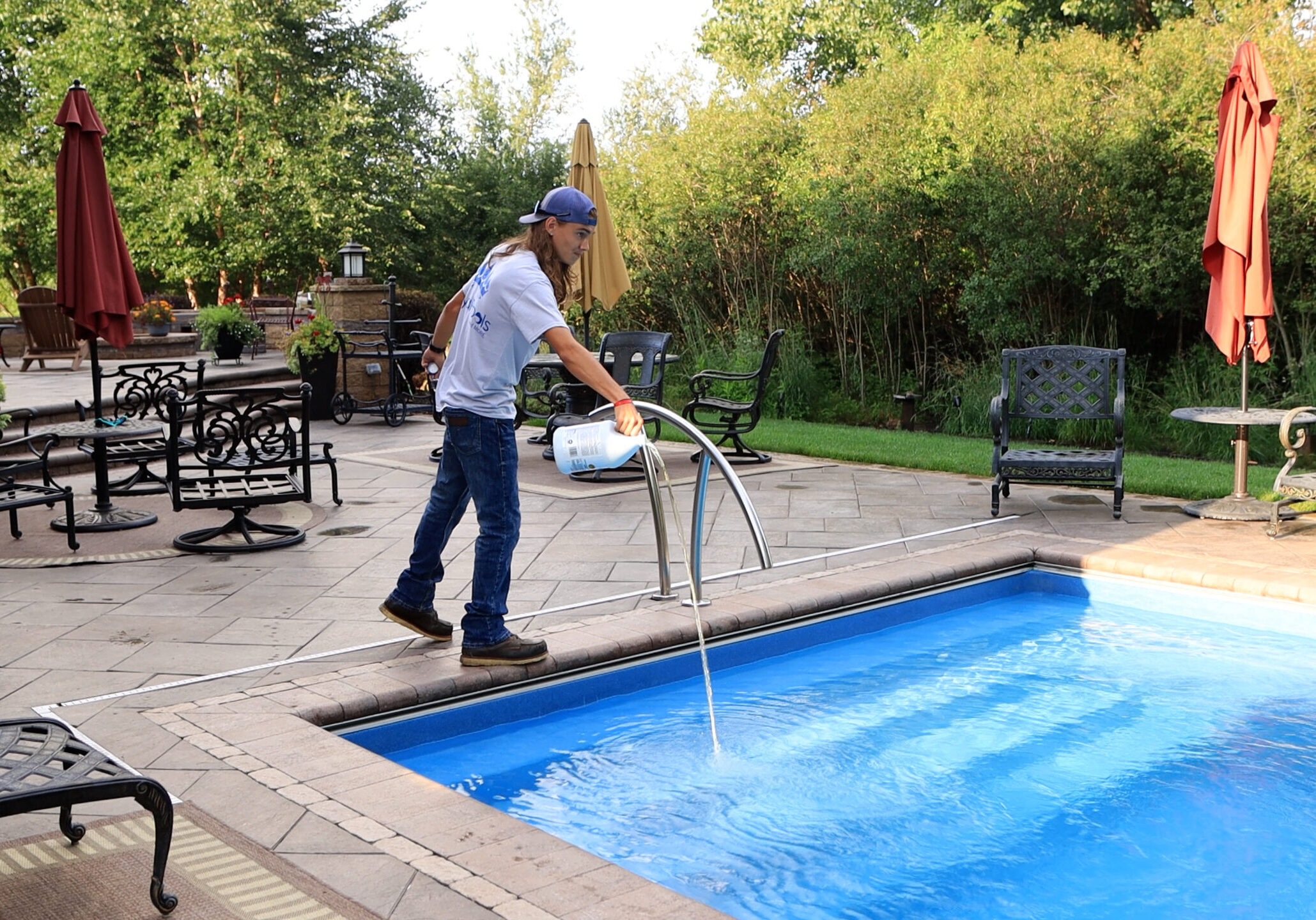 Photo of a man standing over the shallow end of a light-blue inground pool pouring a yellow liquid into the pool. Behind the pool is a stamped concrete patio with chairs and a retaining wall. Trees can be seen behind the patio.