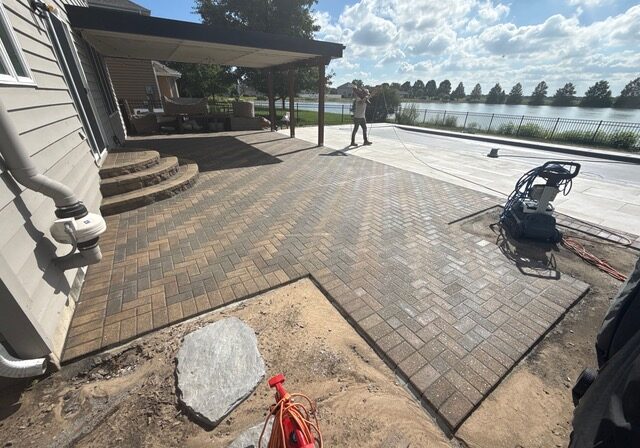 Partially complete brick paver patio off the back of a house with a covered inground pool. In the background is a pond with trees behind it.