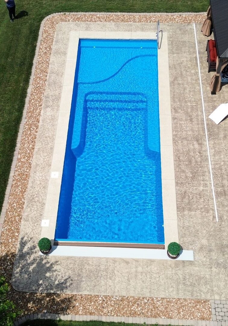 Overhead view of a light-blue inground pool with a light-gray concrete border surrounded by a larger stamped concrete patio connected to a house. Atop the patio is a gazebo near the right-side shallow end corner and a wood deck that extends off the house. Grass can be seen next to the patio.