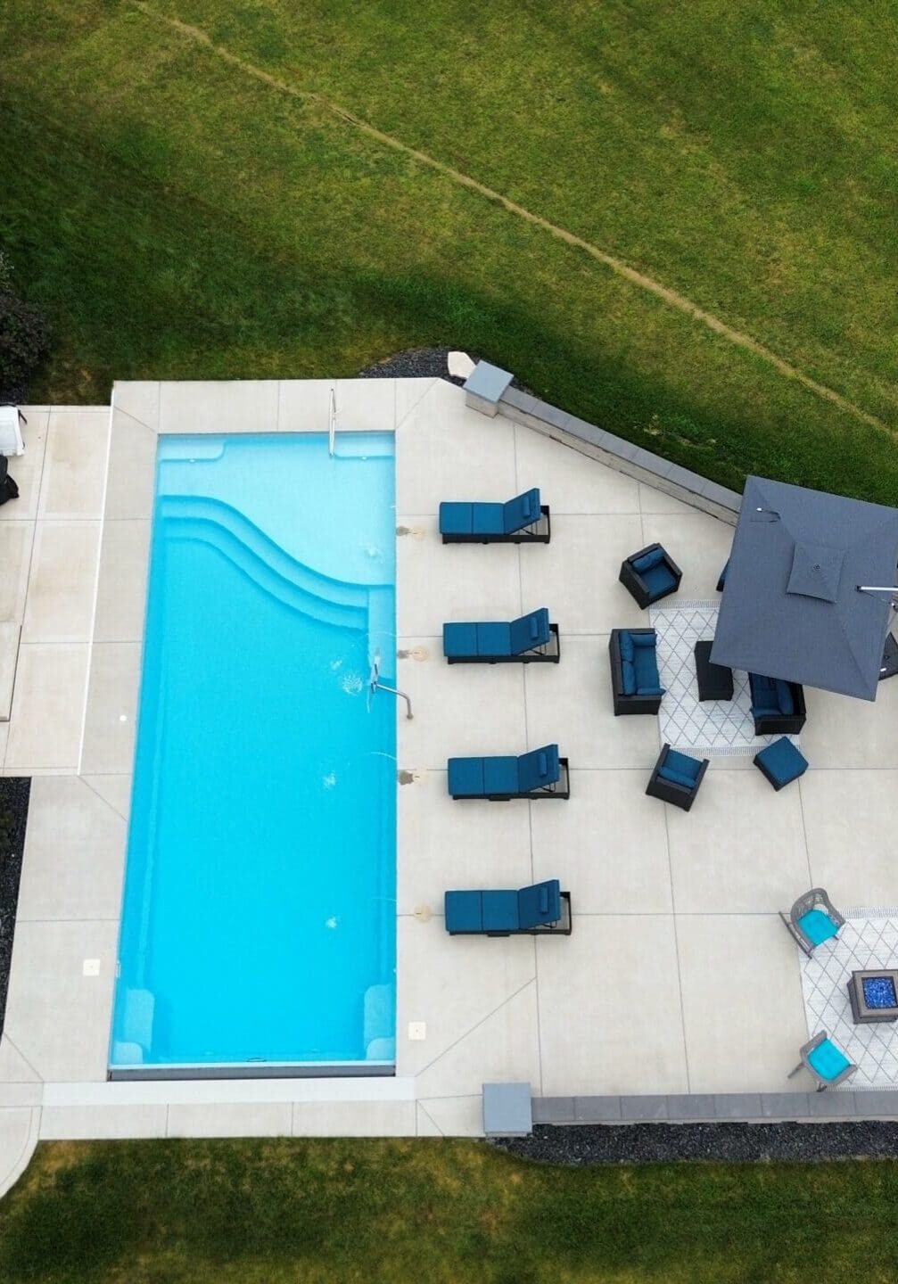 Aerial overview of a backyard featuring a light-blue inground pool surrounded by a large concrete pool deck with a retaining wall on the outside bottom-left corner. Between the pool and the retaining wall are several lounge chairs as well as an umbrella.