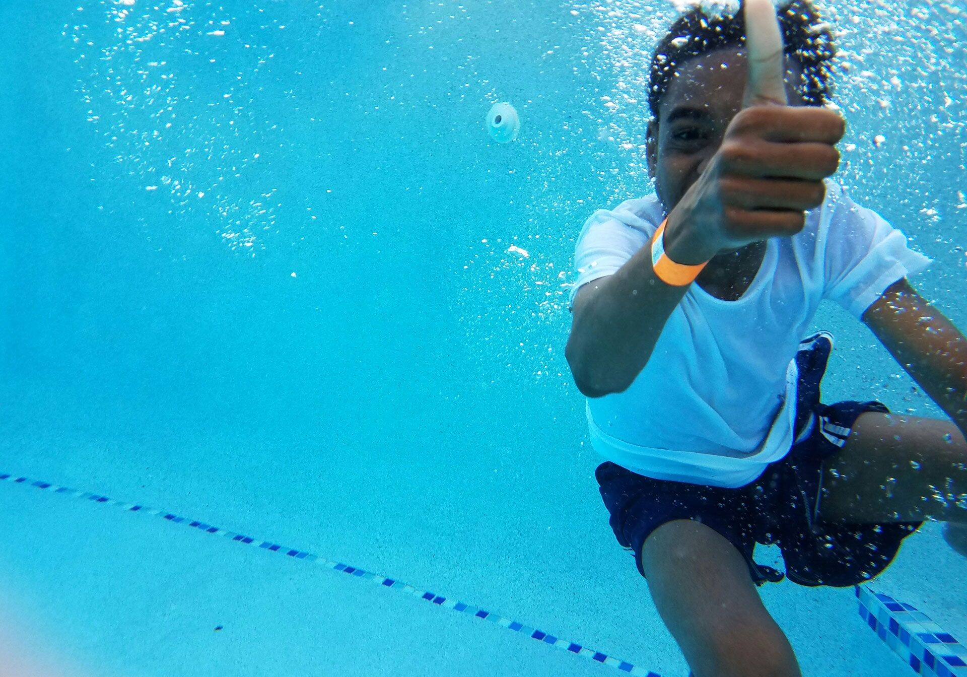 Photo taken of a young boy swimming in a pool. The young boy is holding a thumbs up to the camera.