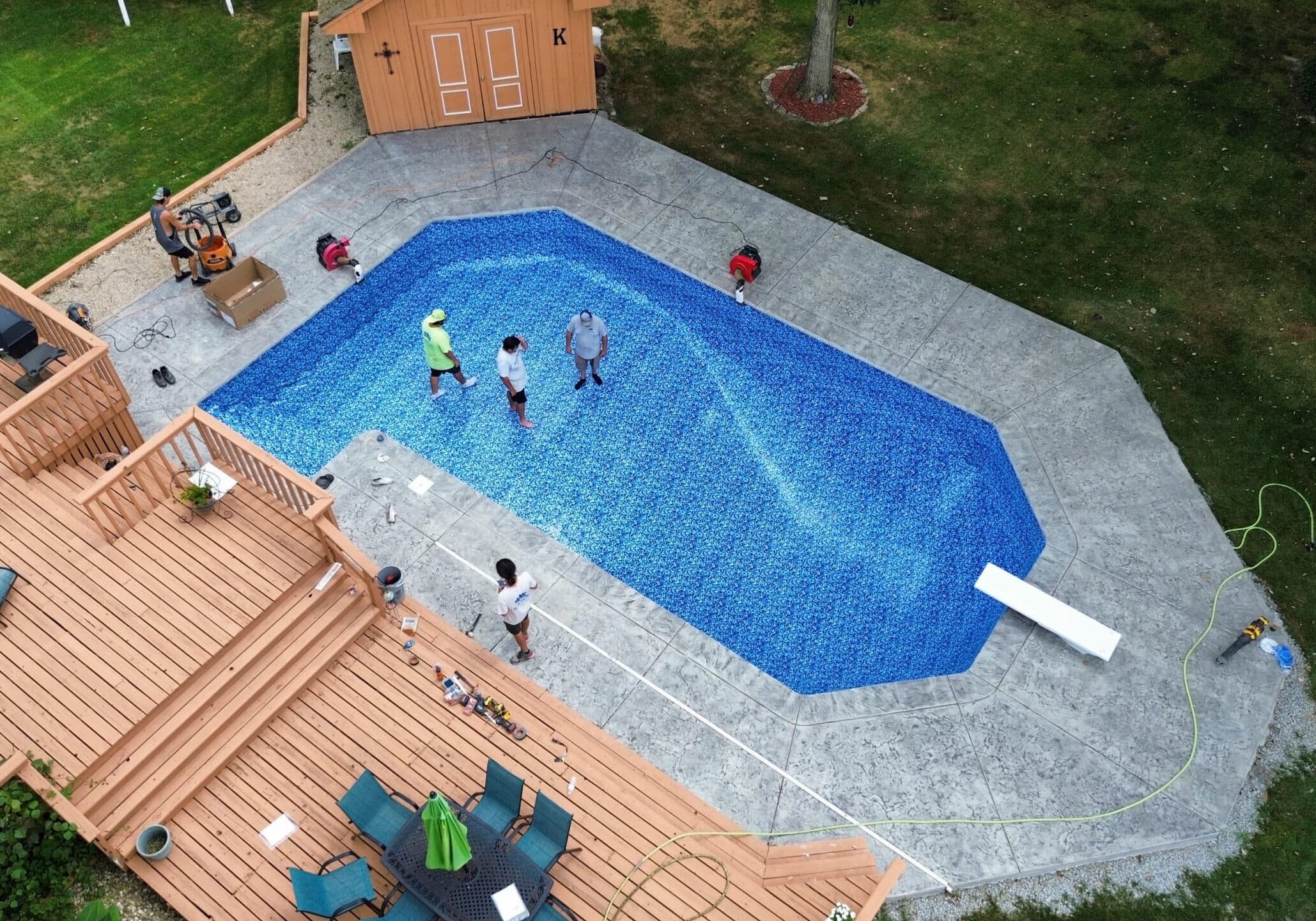Aerial view of a backyard with an empty vinyl liner inground pool with a diving board surrounded by a large gray concrete patio. off the top left corner of the patio is a light-brown shed. Under the pool is a large light-brown wood deck with a black table and multiple blue chairs and tanning chairs. Three men stand in the shallow end of the pool and one stands on the patio near the middle of the wood deck.