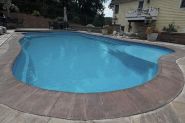 Landscape photo of a freeform inground fiberglass pool surrounded by a brick paver border, with a larger concrete patio surrounding the pool. Next to the pool in the upper right corner of the image. Behind the pool and house in the background are large trees, blocking most of the sky.
