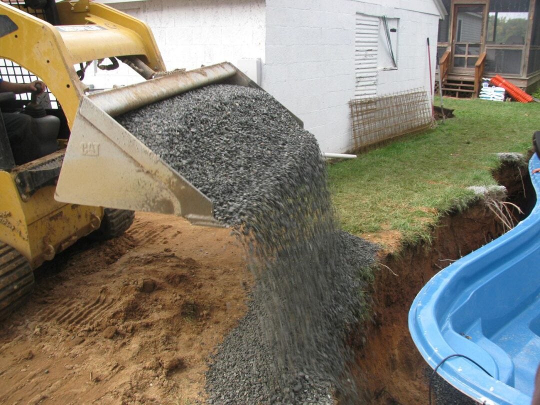 An excavator dumping crushed stone into a hole around a fiberglass pool.