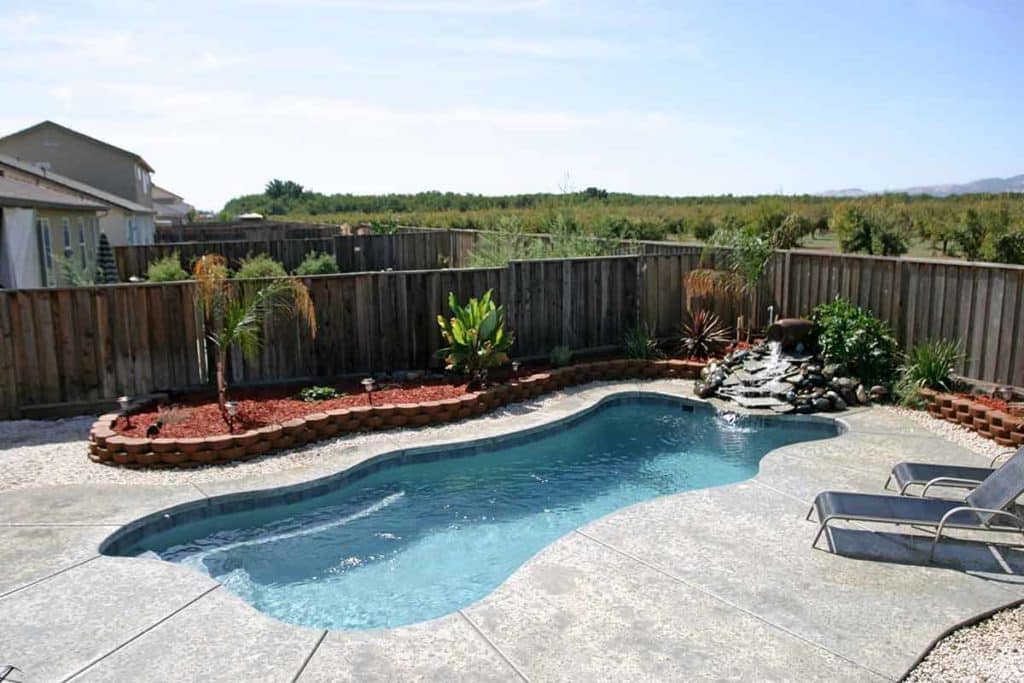 Picture of a backyard with a freeform inground pool. The pool is surrounded by gray stamped concrete and surrounded by rock and a raised mulch bed with plants. The entire area is surrounded by a wood fence. Trees and other homes can be seen beyond the yard.