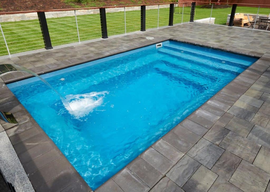 Light-blue rectangular inground fiberglass pool surrounded by dark-gray concrete pavers surrounded by a fence. The pool features a spillover pouring water into the deep-end on the far left-side of the image.. Beyond the fence in the background is grass and a retaining wall on the outside. 