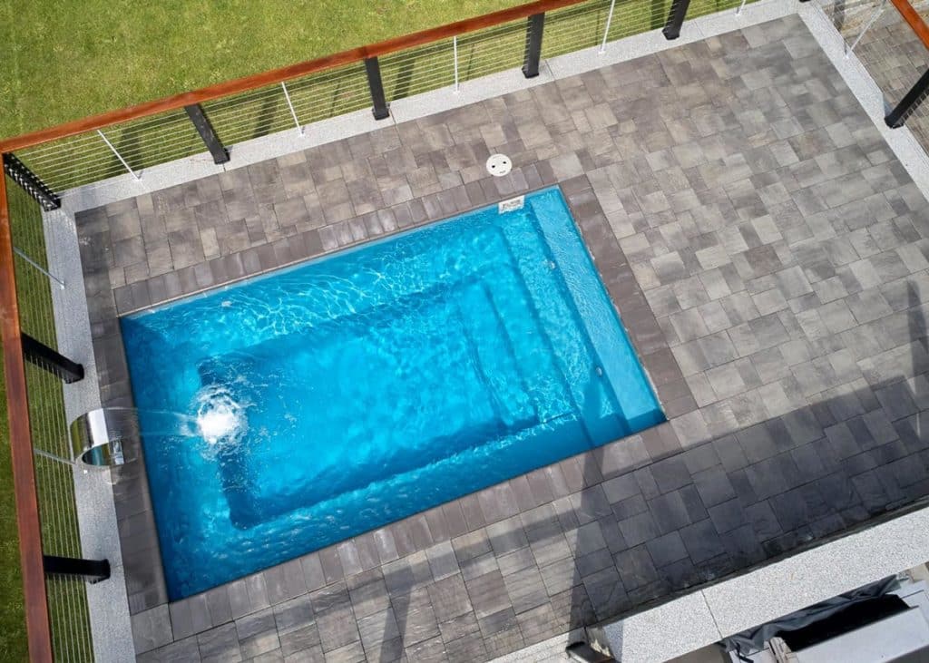 Aerial landscape photo of a light blue, rectangular inground pool surrounded by a brick paver patio, which is enclosed by a steel and wood fence. The pool features a waterfall spillover in the deep end close to the bottom left corner of the image. The pavers are gray and square-shaped. Outside the patio beyond the fence is green grass. 