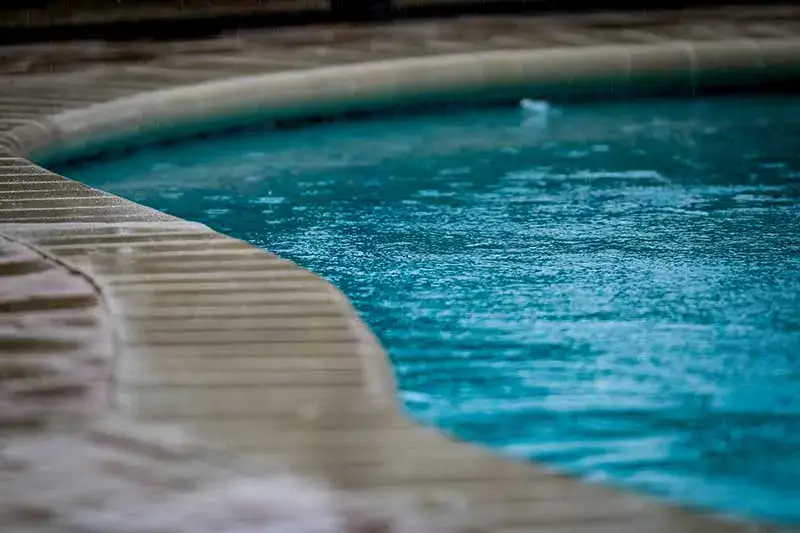 A closeup image of part of a freeform inground pool. Rainfall is getting into the pool and the concrete is wet.